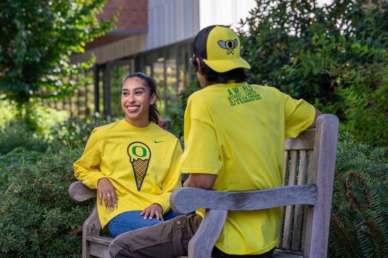 Two people sitting on a bench wearing yellow shirts. One person is facing toward the camera but looking off to the side while the other has their back to the camera. Both are wearing apparel from the Flight and Fight collection.