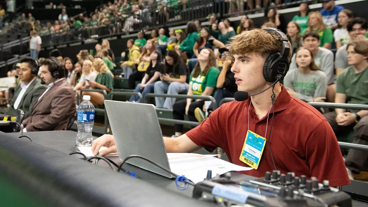 A UO student sitting at a table for a sports broadcast with a crowd of Oregon students and fans in the background