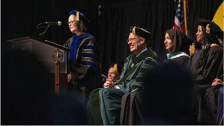 UO President John Karl Sholz is seated amongst UO faculty members at his investiture ceremony. To his right, Karen Ford, interim provost and senior vice president, is standing at the podium, delivering remarks. All on stage are wearing full academic regalia.