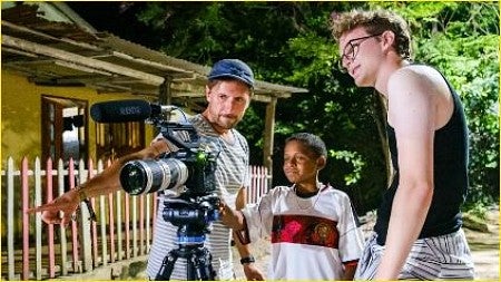 Outside a house in Colombia, a young boy looks through the camera viewfinder as two SOJC students line up a shot.